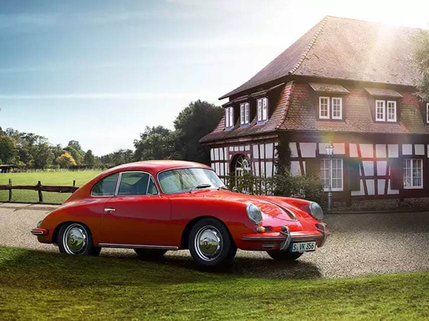 A classic red car parked on grass near a charming half-timbered house under a sunny sky.